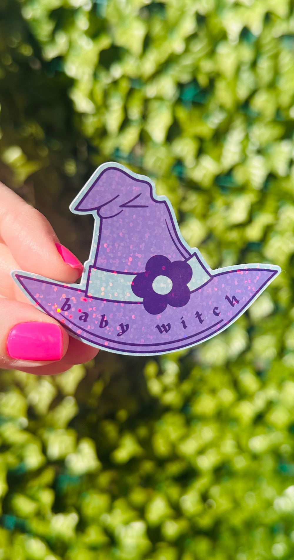 womans hand holding a purple witches hat sticker that says baby witch