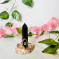 black obsidian tower sits upon a redish petrified base handcrafted into a crystal ring holder. sitting in a white background with greenery and florals. ring holder fits ring sizes 4 and larger