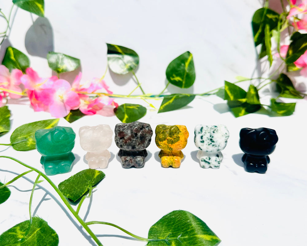 hello kitty crystal carvings. sitting on white marble surrounded by vines and flowers. green aventurine. rose quartz. yooperlite. bumblebee jasper. moss agate. black obsidian.