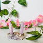 two kunzite towers sit on a pink amethyst base. handcrafted crystal ring holder fits ring sizes 2.5 and bigger. piece sits on white marble surrounded by greenery and flowers