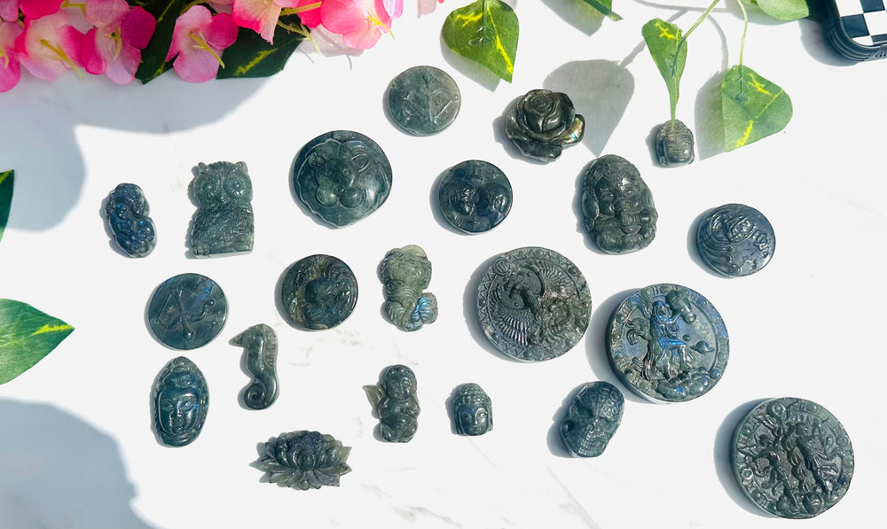 blue flash labradorite coins and carvings sitting on white marble surrounded by greenery and flowers. tigers. owls. budha. lotus flower. seahorse. crab. cancer. leo. zodiac. rose. aquarius. virgo. eagle. cherub. gemini. 