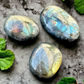 3 beautiful labradorite palm stones sit on concrete showing off colorful flashes of blue, green, orange, yellow and more.