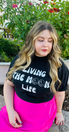 woman wearing an odd squad shirt that says living the moon life, she is sitting outside.