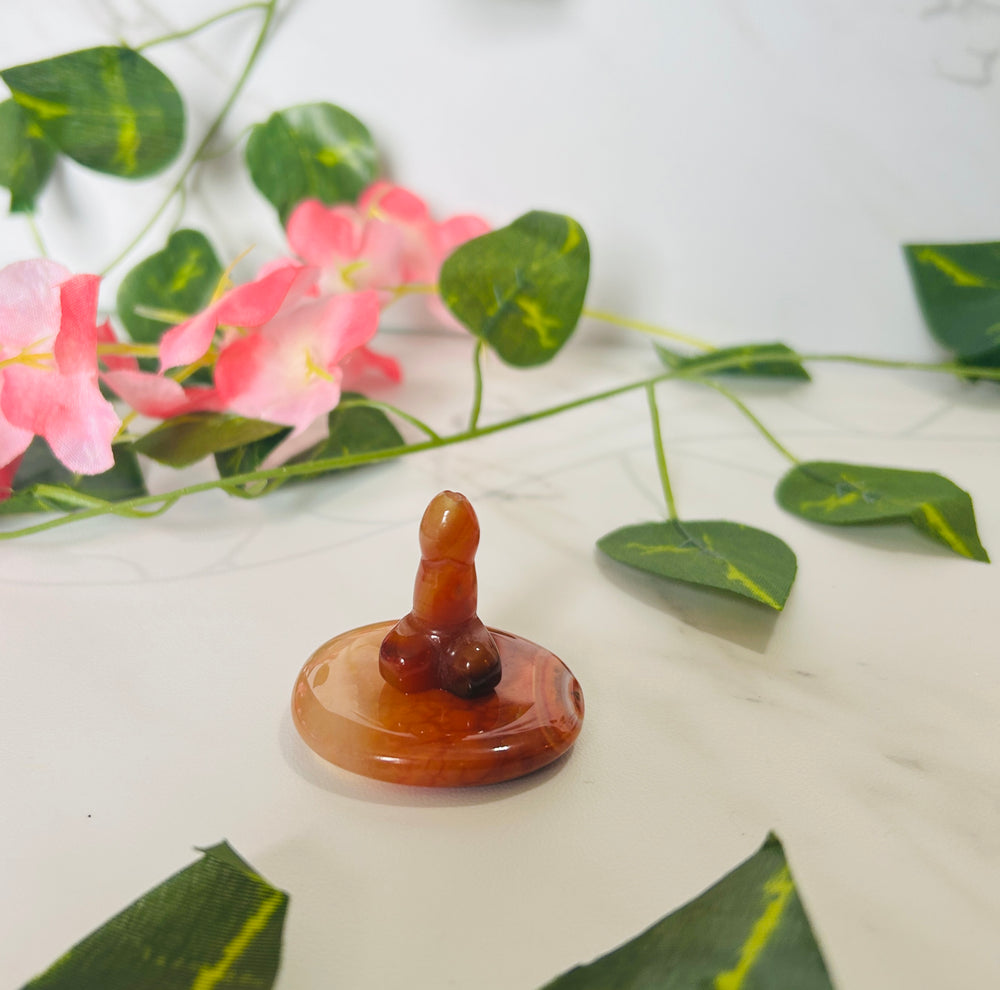 carnelian and agate crystal phallic incense holder on a white counter with leaves and pink flowers