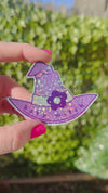 womans hand holding a purple witches hat sticker that says baby witch