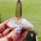a video of smokey quartz crysal tower sitting on a polished blue flower agate base forming a handcrafted crystal ring holder fitting ring sizes 4.5 and bigger in front of a green  field