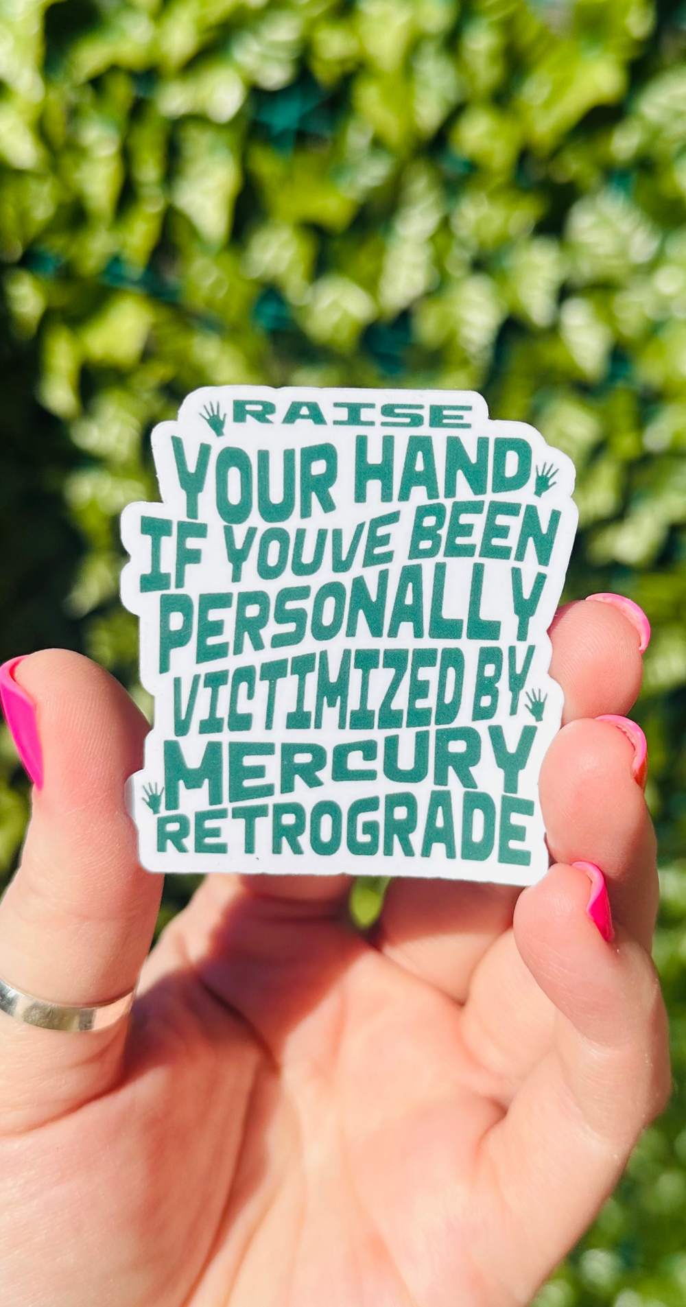 womans hand holding a green and white sticker that says "raise your hand if you've been personally victimized by mercury retrograde"