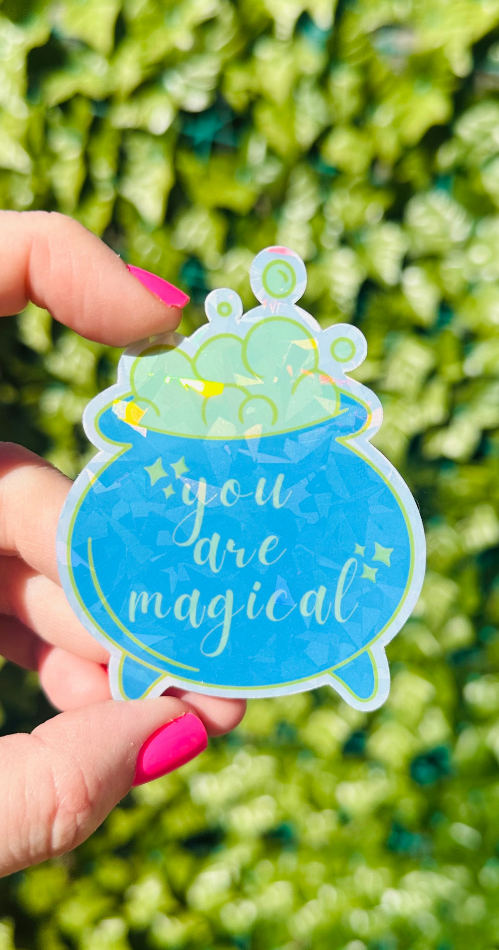 a woman holding a green and blue cauldron sticker that says you are magical.